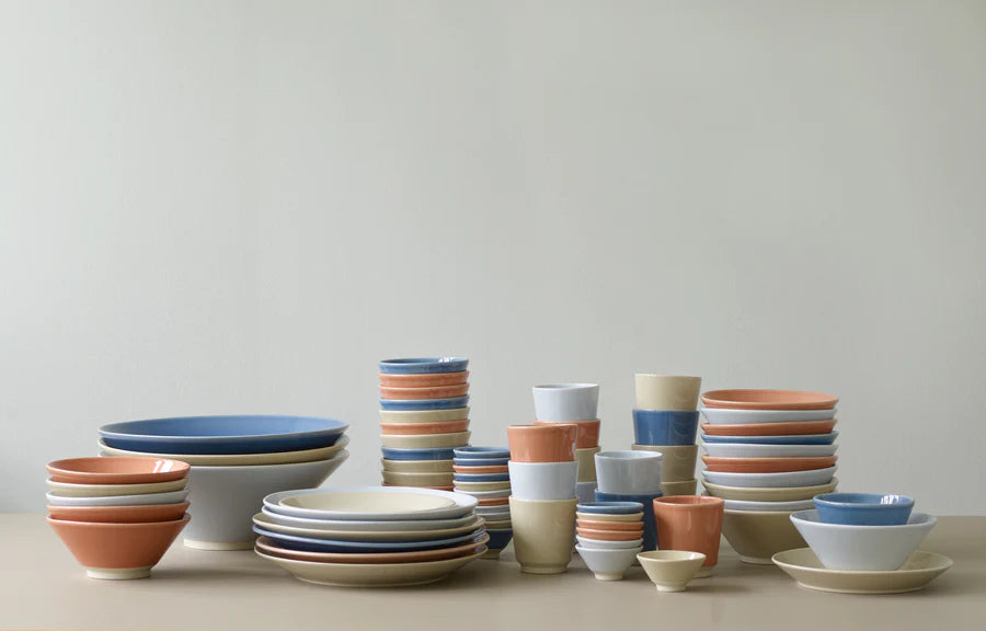 Stakke af keramikservice i forskellige størrelser og farver står ordnet på en beige overflade mod en neutral væg. Farverne inkluderer blå, orange, hvid og beige.