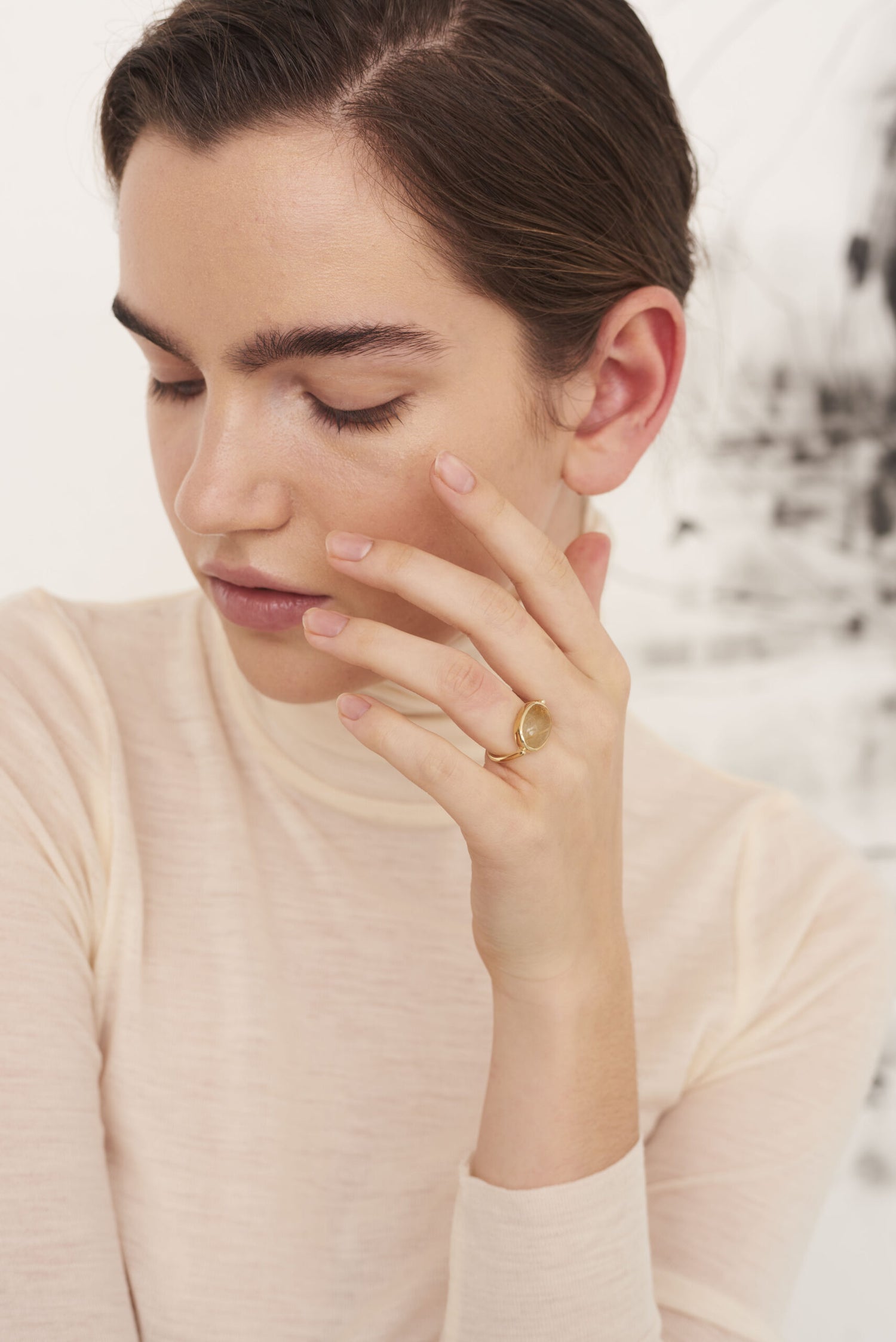 En kvinde med tilbagetrukket brunt hår hviler sin hånd ved sit ansigt, hvor en guldring med en rund sten kan ses. Baggrunden er lys med et abstrakt maleri.