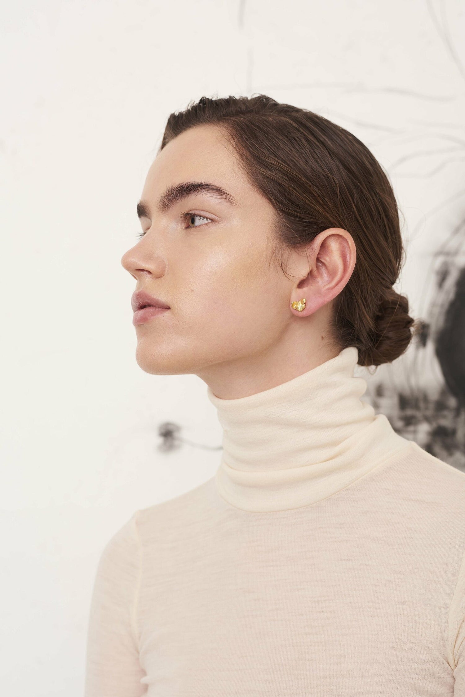 En kvinde ser til venstre, iført en rullekrave og små guldøreringe. Hun står foran en lys baggrund med en abstrakt tegning.