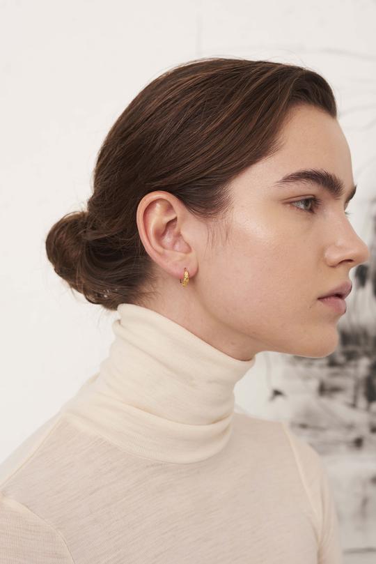 En kvinde med brunt hår sat op, bærer en lille guld ørering i sit venstre øre. Hun er iført en cremefarvet rullekrave.