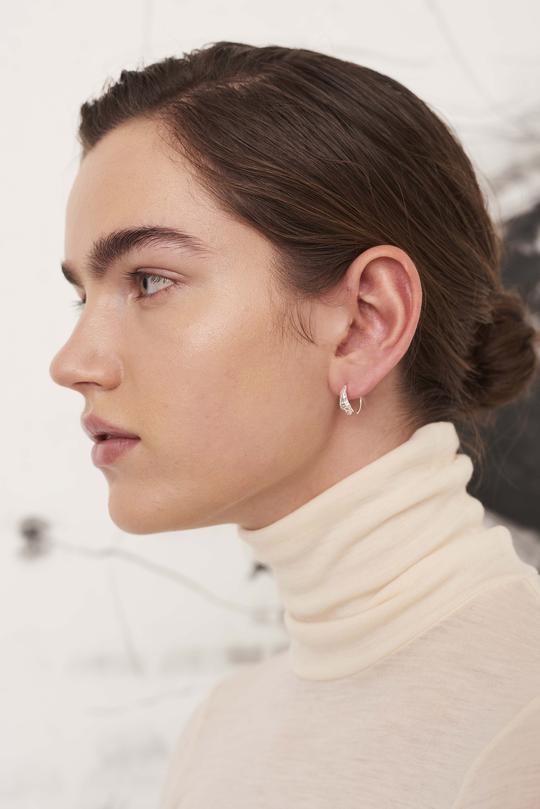 En kvinde viser et sølvfarvet ørering i øret, mens hun poserer med en cremefarvet rullekrave foran en lys, abstrakt baggrund.