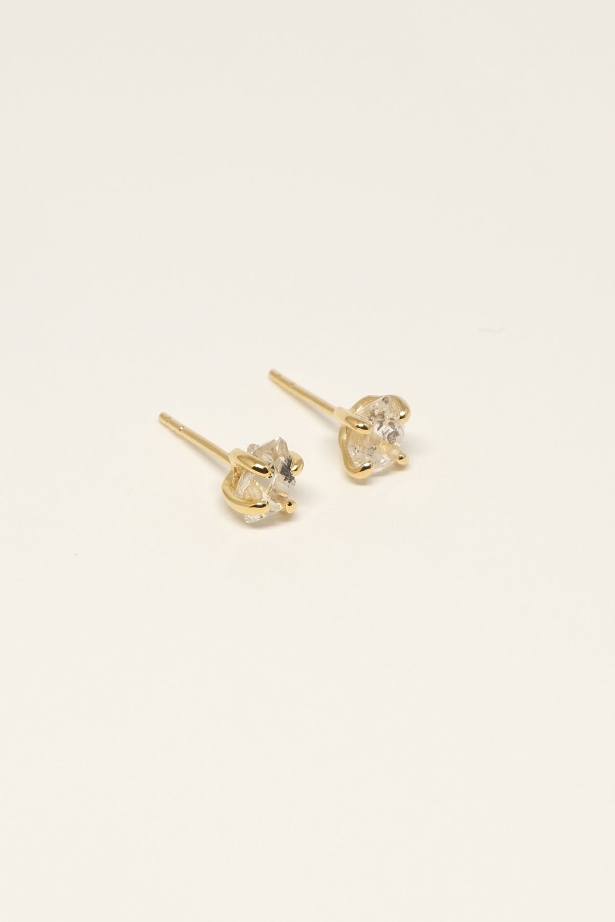 To gyldne øreringe med uregelmæssigt formede, klare sten ligger på en ensfarvet, off-white overflade.