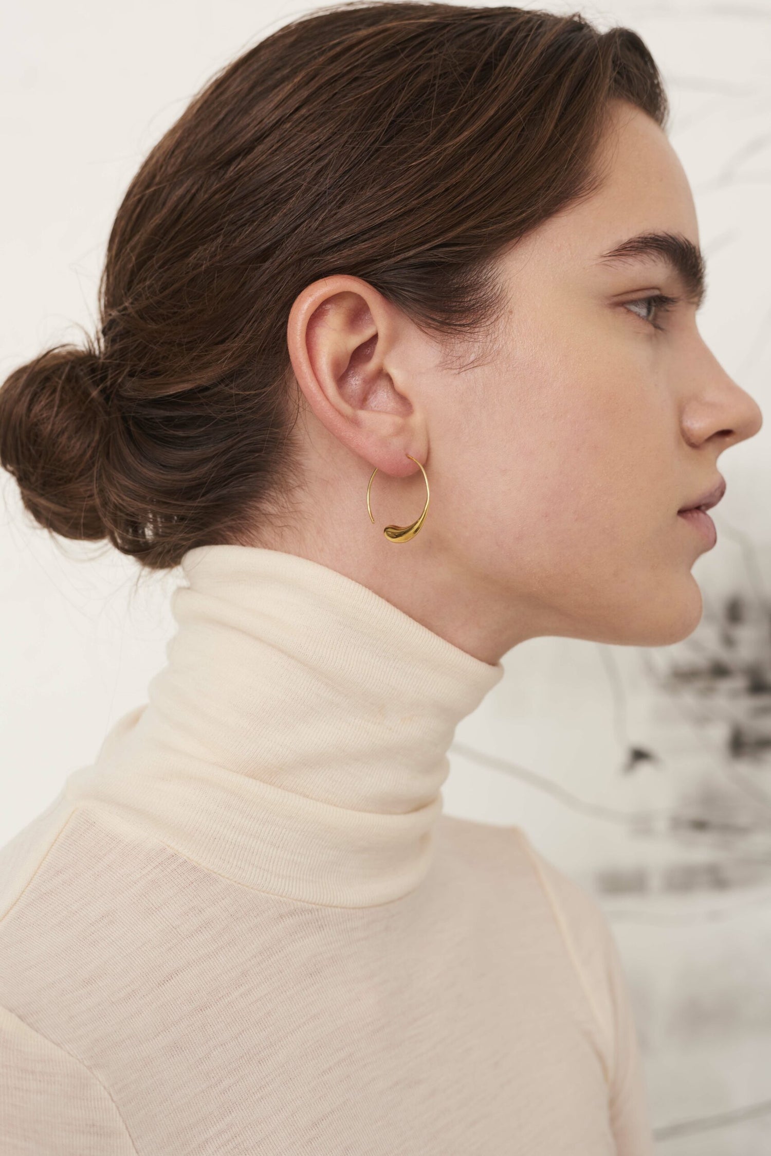 En kvinde med opsat hår har en guldfarvet dråbeformet ørering i øret, og hun bærer en cremefarvet rullekrave i et lyst miljø.