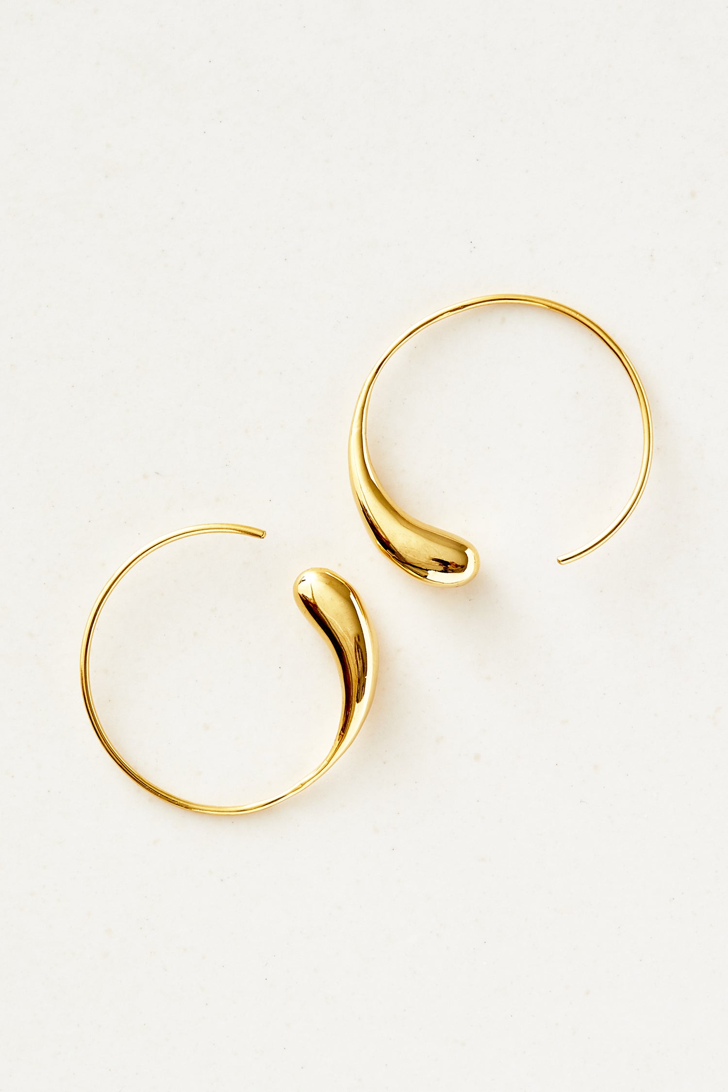 To guldøreringe, formet som åbne cirkler med en dråbeformet detalje, ligger på en hvid overflade.