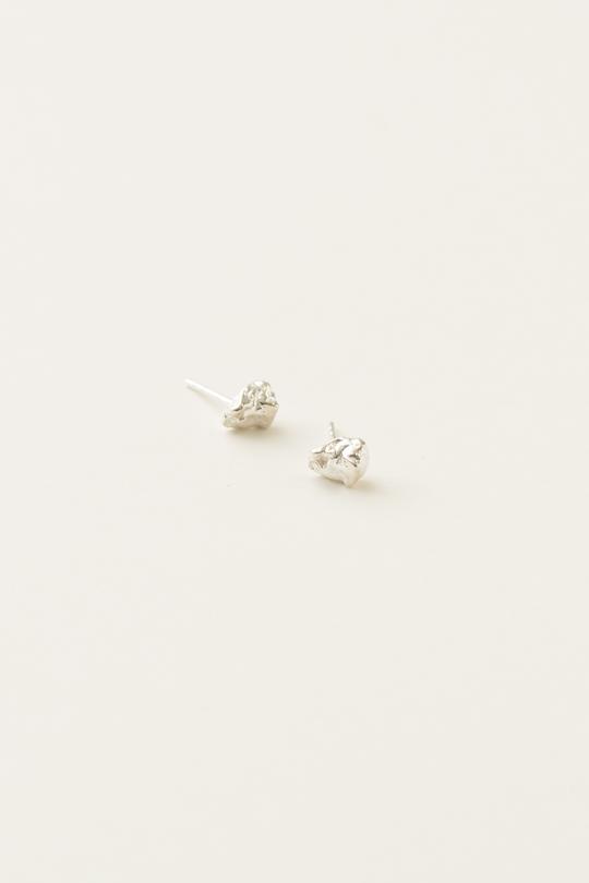 To sølvfarvede øreringe ligger på en hvid overflade. Øreringene er af ujævn form og har en stift til at fastgøre dem.