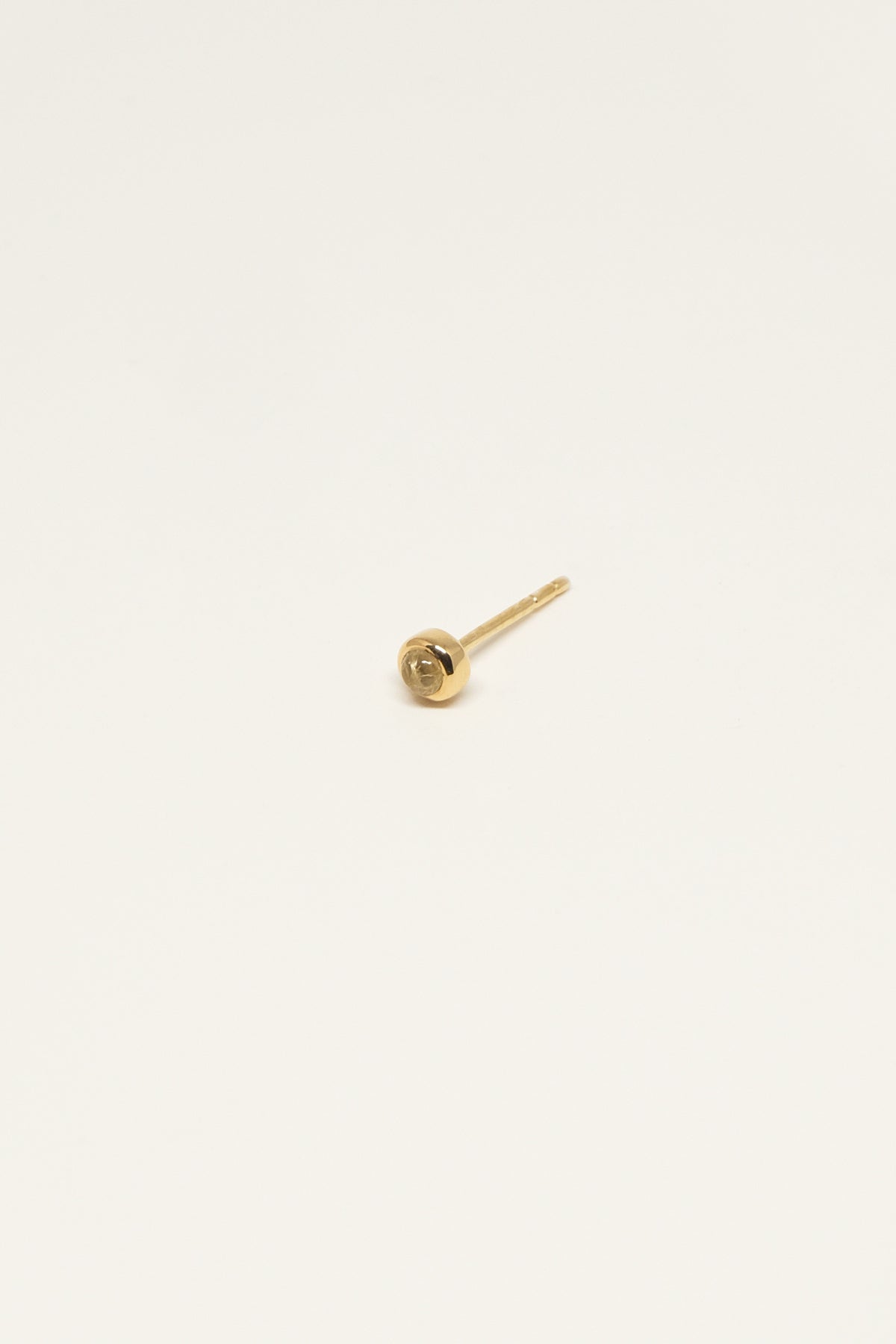 En enkelt guldørering ligger på en ren, lys creme baggrund. Øreringen består af en lille, rund fatning og en lige stift.