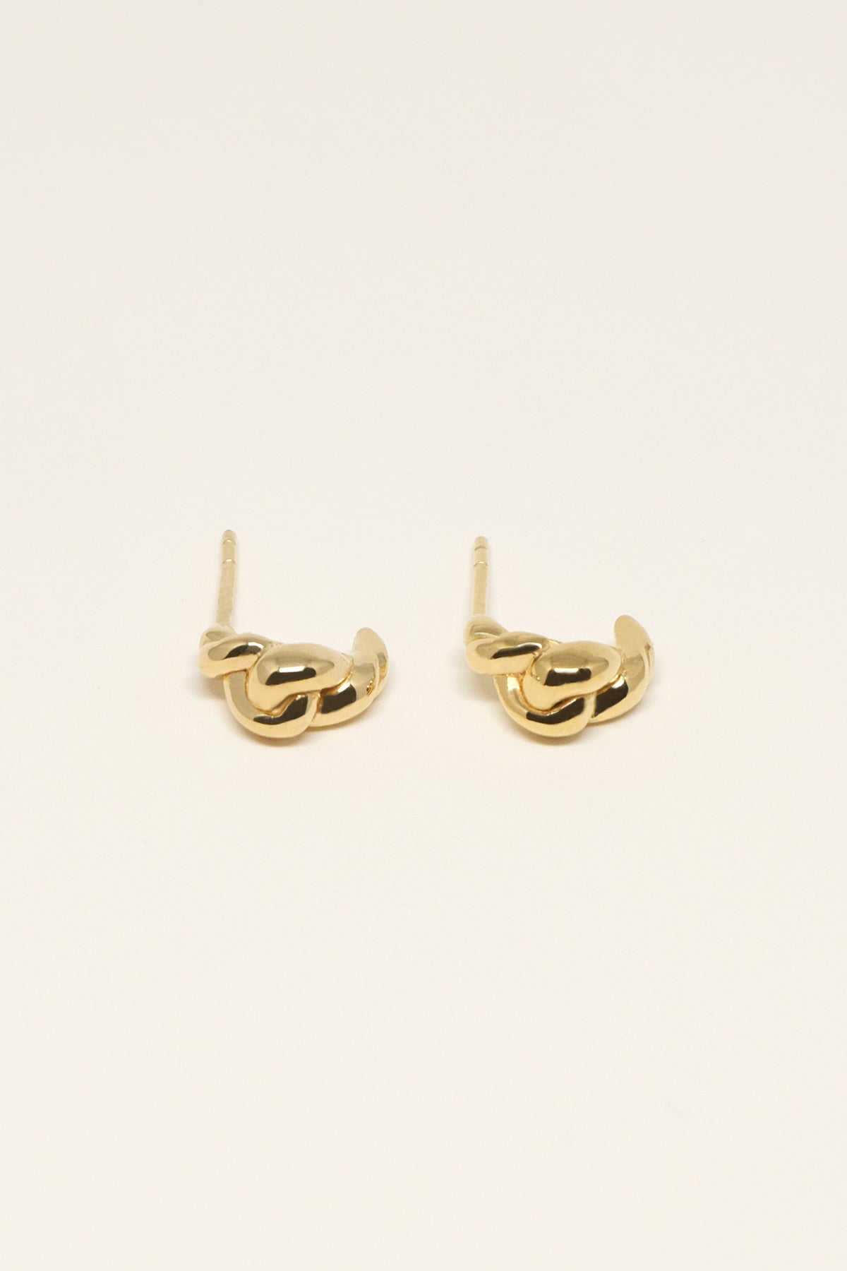 To gyldne øreringe, formet som sammenflettede led, sidder på en lys beige overflade. Øreringene har en studs, der stikker op fra det sammenflettede design.