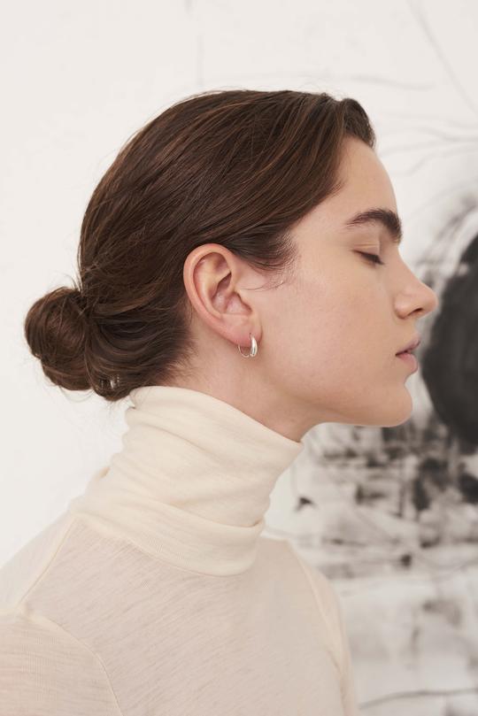 En kvinde med opadgående hår bærer en sølvfarvet ørering, mens hun kigger mod højre. Hendes baggrund er hvid med abstrakt kunst.