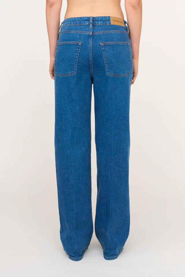Person står i blå jeans med brede ben, set bagfra mod en hvid baggrund. "Tomorrow" står på etiket.
