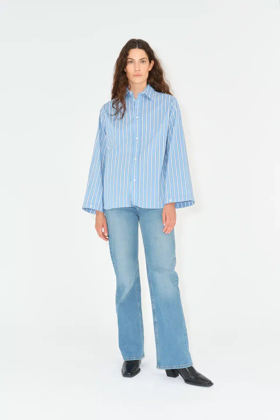 En kvinde står, iført en blåstribet skjorte, jeans og sorte sko. Hun kigger direkte fremad i en hvid baggrund.
