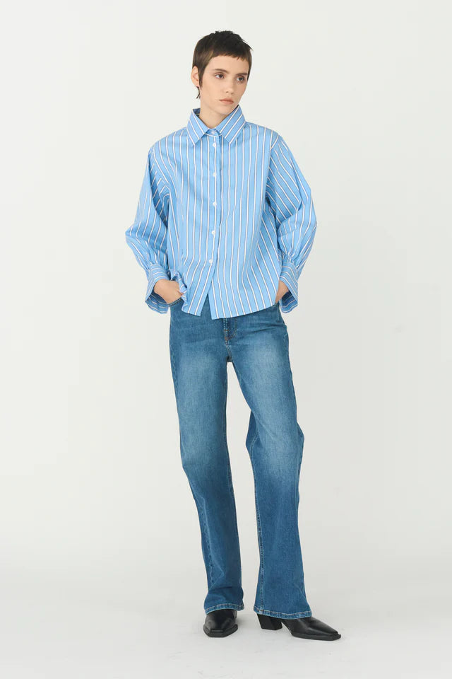 En model står med hænderne i lommerne, iført en lyseblå stribet skjorte og jeans, i et lyst studie.