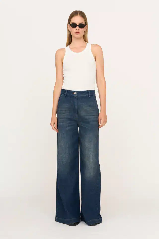 En kvinde poserer stående iført en hvid tanktop og brede, blå jeans foran en hvid baggrund. Hun har solbriller på.