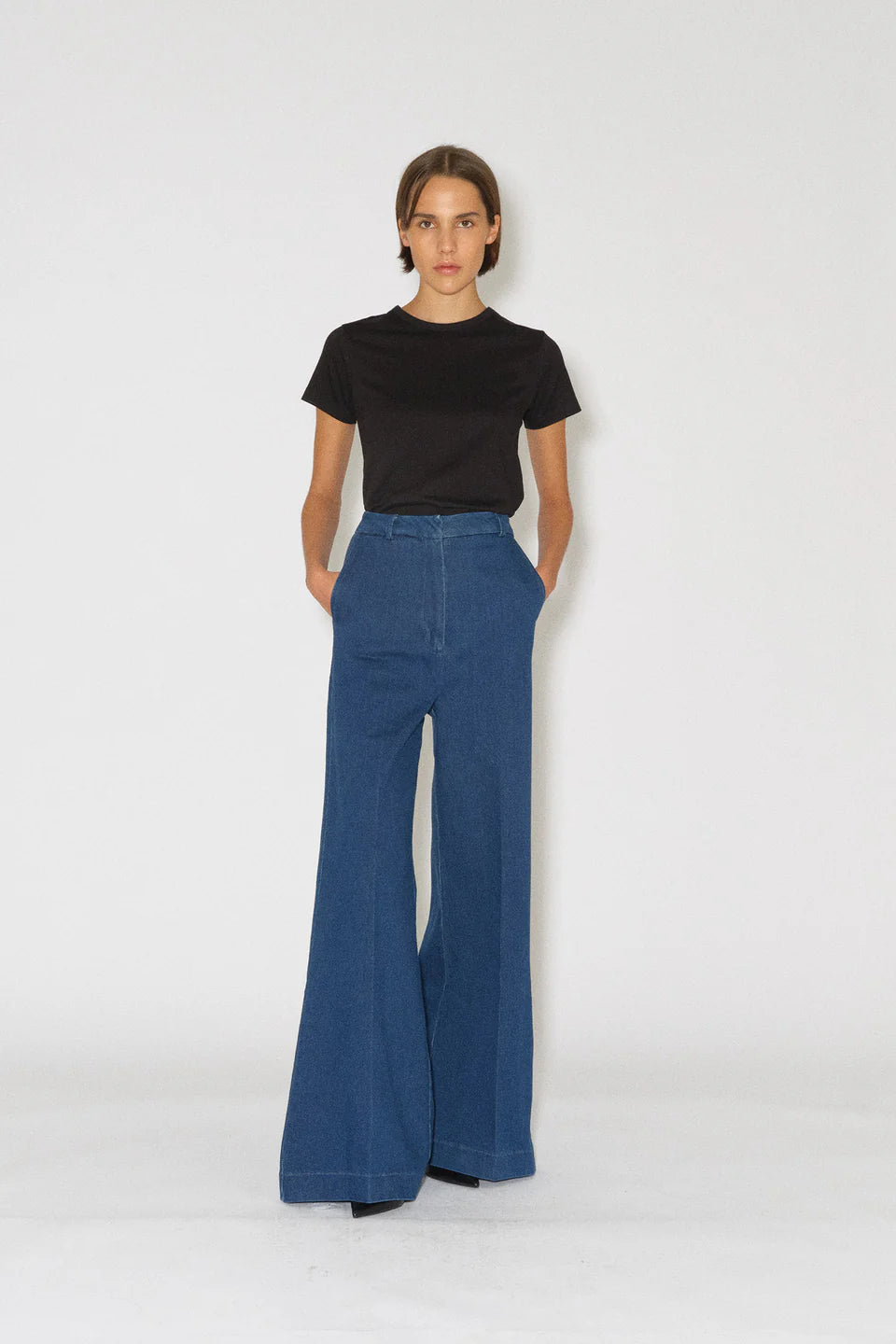 En person står med hænderne i lommerne, iført en sort t-shirt og blå denim bukser med høj talje og vide ben, mod en hvid baggrund.
