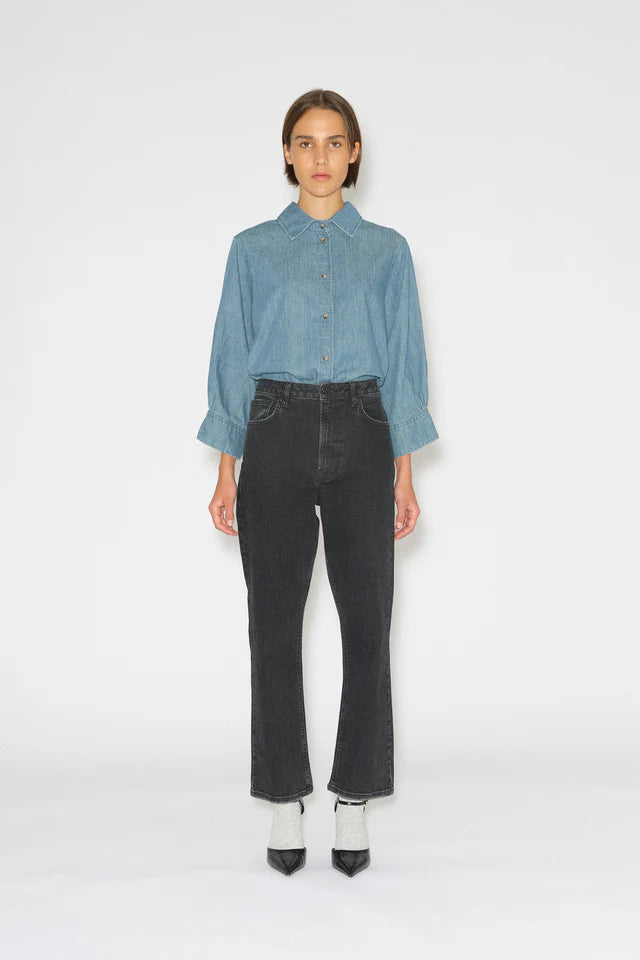 En kvinde står i et hvidt miljø, iført en lyseblå skjorte med lange ærmer, højtaljede sorte jeans og sorte stilethæle med hvide sokker.
