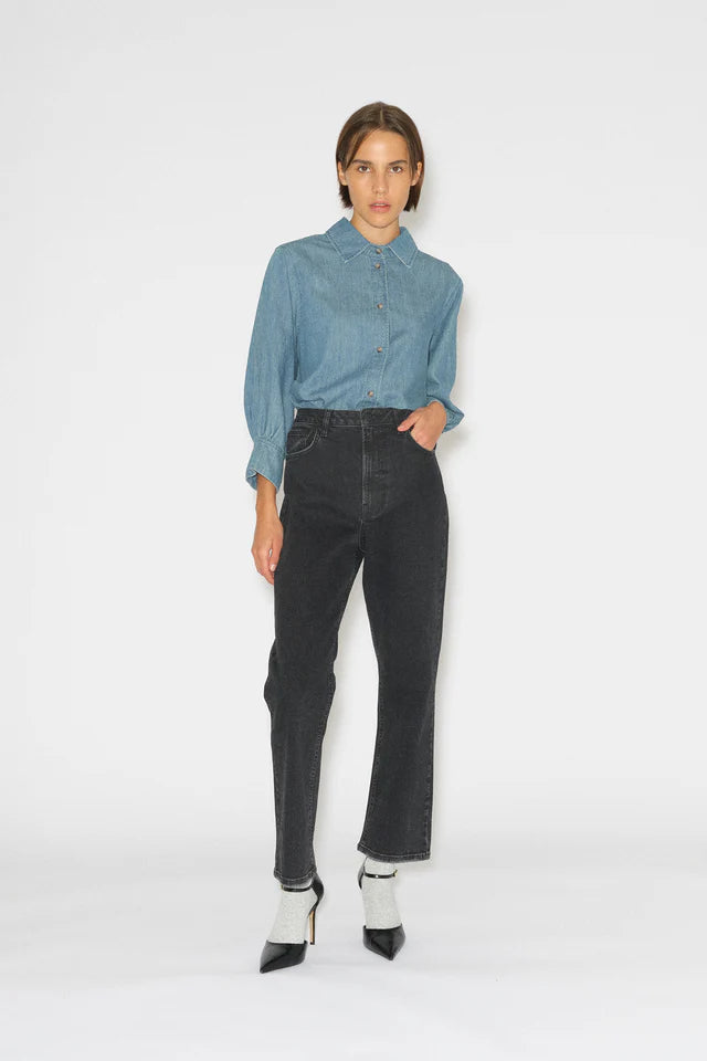 En kvinde står, med en hånd i lommen, iført en denimskjorte, sorte jeans og sorte højhælede sko med glitterstrømper på en hvid baggrund.