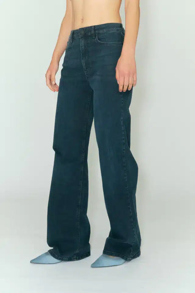 Personen står i brede, mørkeblå jeans og lyseblå sko foran en hvid baggrund.