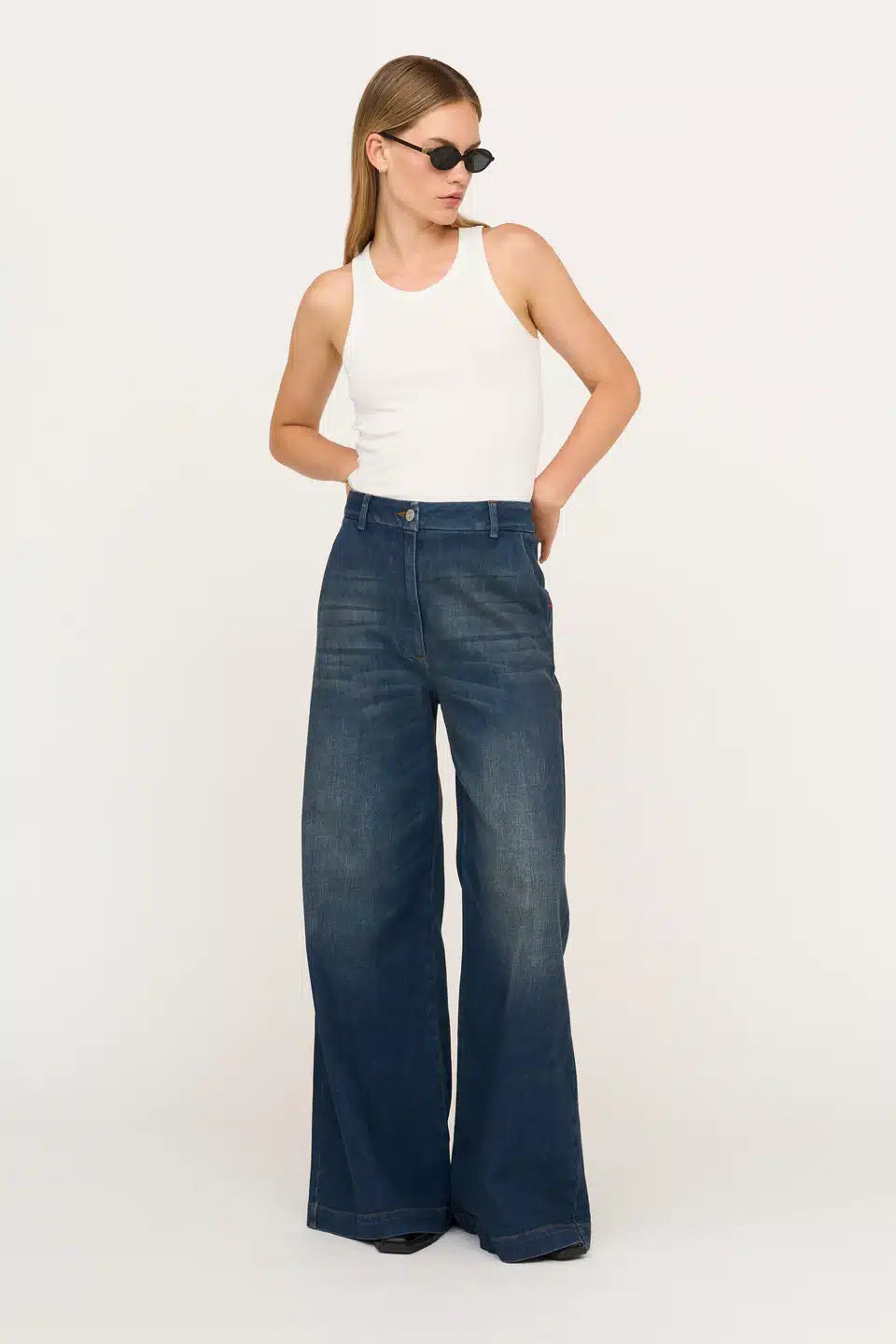En kvinde står, iført en hvid tanktop og mørke denim bukser med vide ben, og holder hænderne i siden foran en hvid baggrund. Hun bærer solbriller.