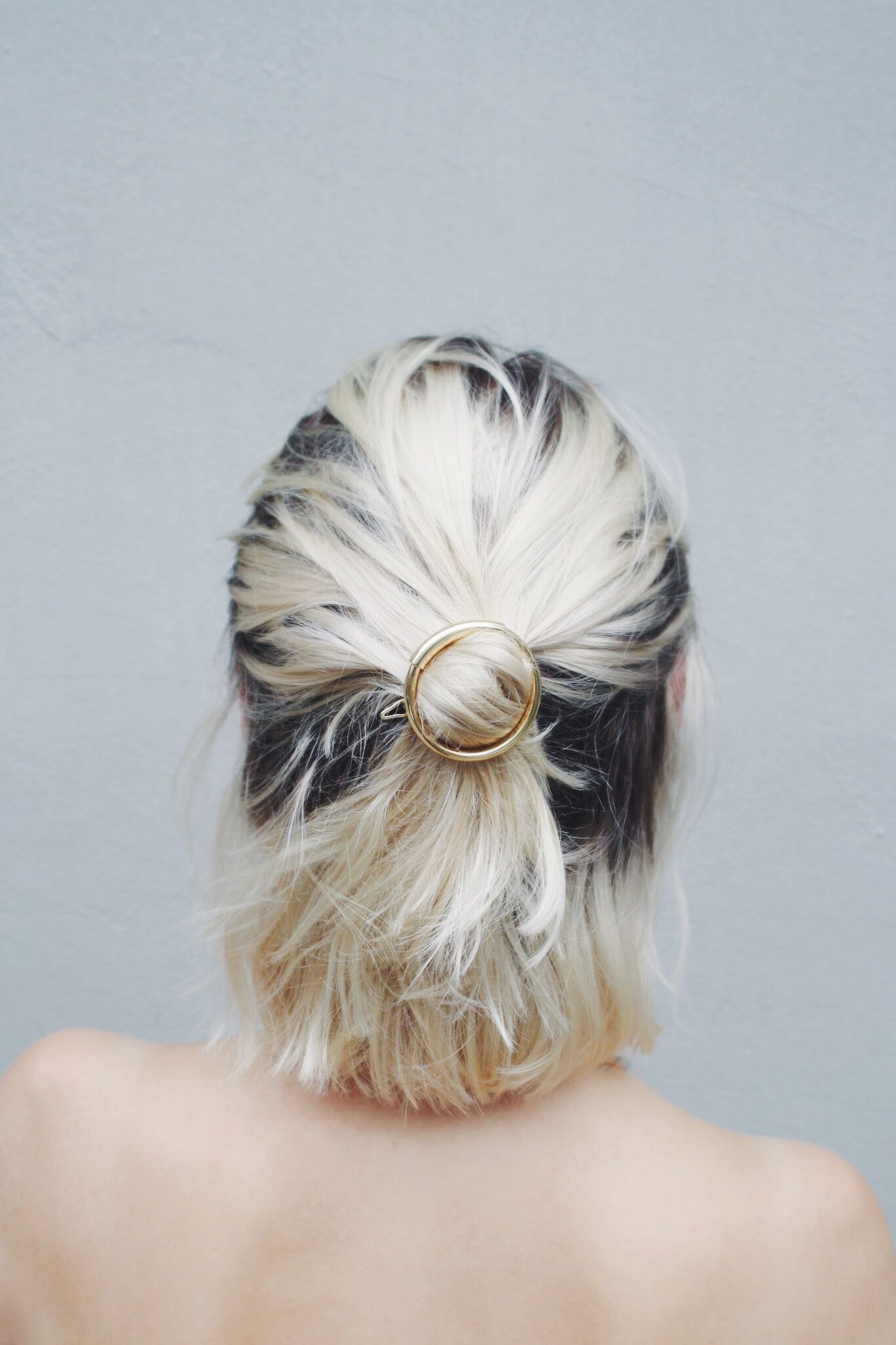 En kvindes nakke og skulderblade er synlige, hendes blonde hår er samlet i en knold med en gylden cirkelspænde, foran en lysegrå baggrund.