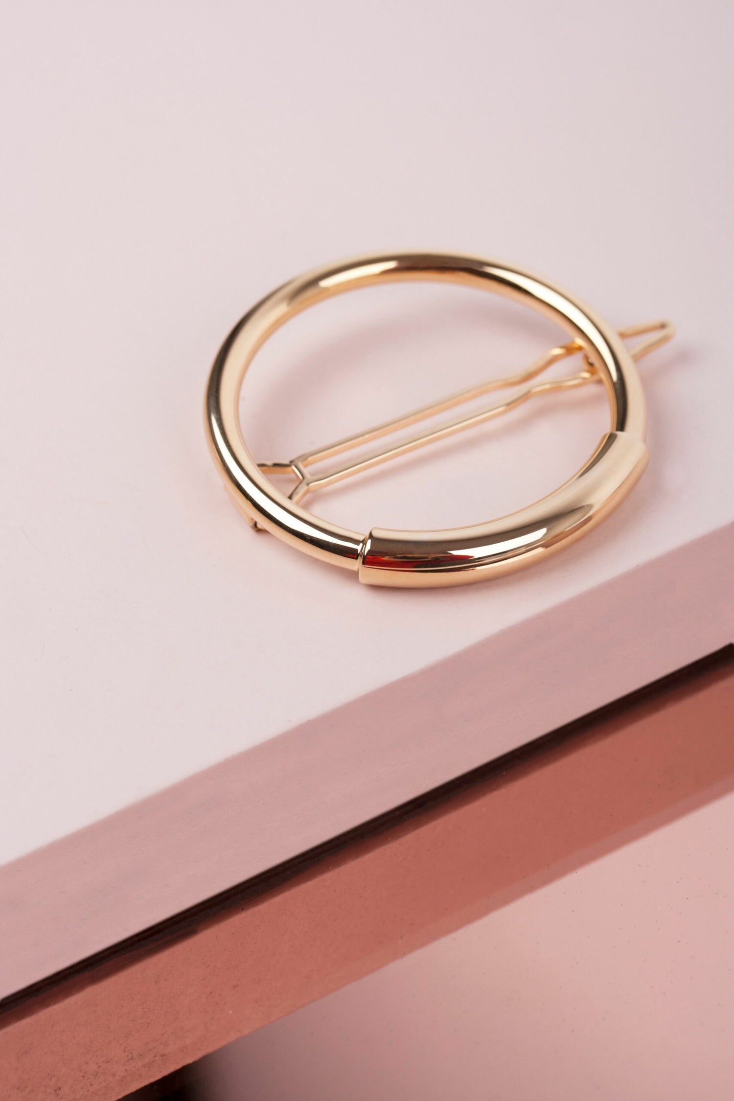 En gylden, cirkelformet hårklemme hviler på en overflade, der er opdelt i lyserøde og rosaguld segmenter.