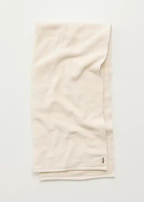 Et cremefarvet håndklæde er foldet og ligger fladt mod en hvid baggrund. Etiketten "IHAJU" er synlig nederst til højre.