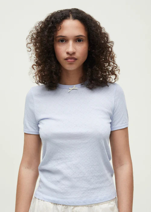 En kvinde med krøllet hår poserer i en lyseblå t-shirt med en lille sløjfe. Hun står foran en neutral, hvid baggrund.
