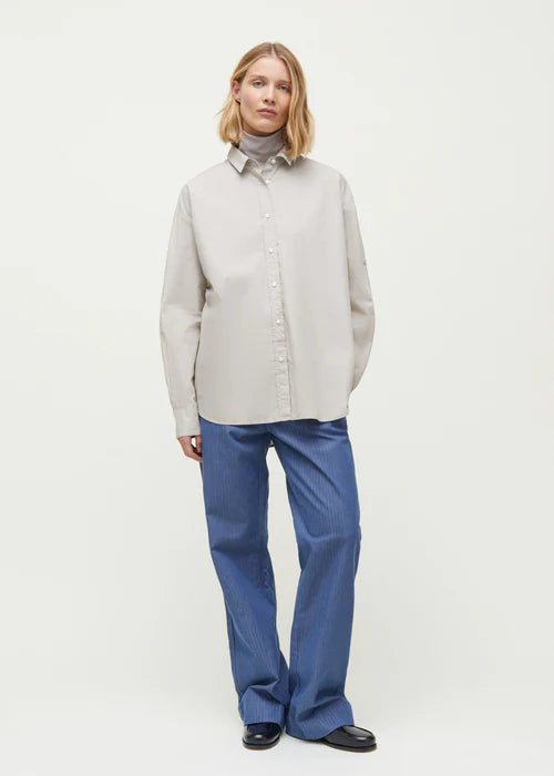 En kvinde står mod en hvid baggrund, iført en lysegrå skjorte, blå bukser, en grå turtleneck og sorte sko. Hun har blonde hår.