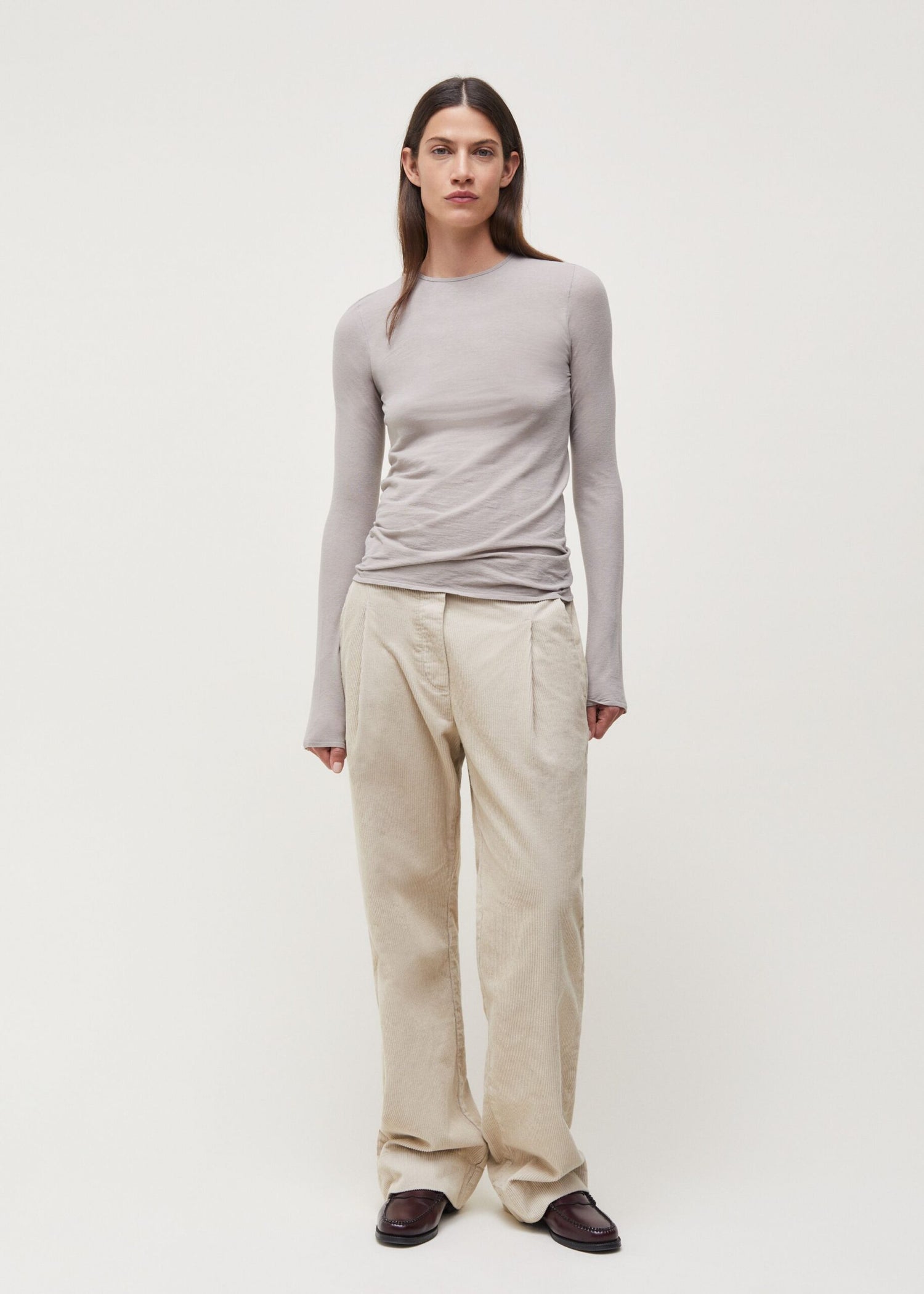 En kvinde står, iført en lysegrå langærmet bluse og brede beige bukser med plissering, på en neutral, lys baggrund. Hun kigger direkte mod beskueren.