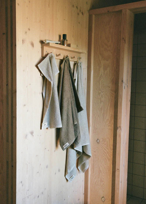 Håndklæder hænger på en træhylde med knager på en trævæg i et badeværelse. Håndklæderne har mærket "deha". En døråbning ses delvist i billedet.