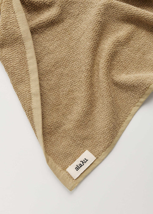 Et beigefarvet håndklæde er foldet, der viser en kant samt et lille mærke med teksten "aïaïu".