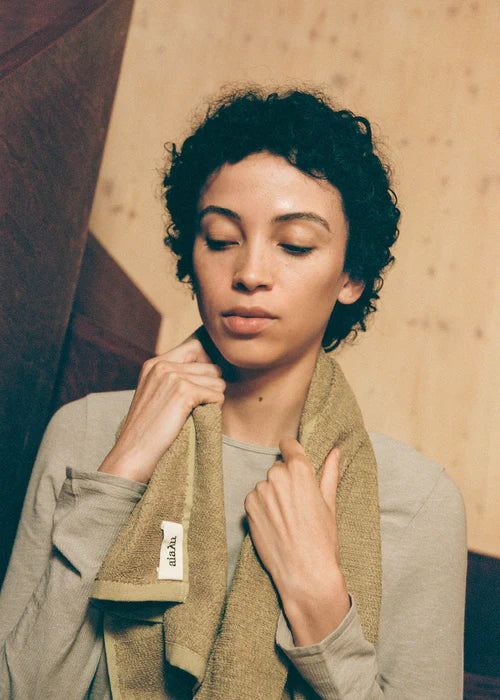 En kvinde holder et olivengrønt håndklæde med "ala.Lu" etiket om halsen. Hun står foran en baggrund med beige vægge og en brun træstruktur.