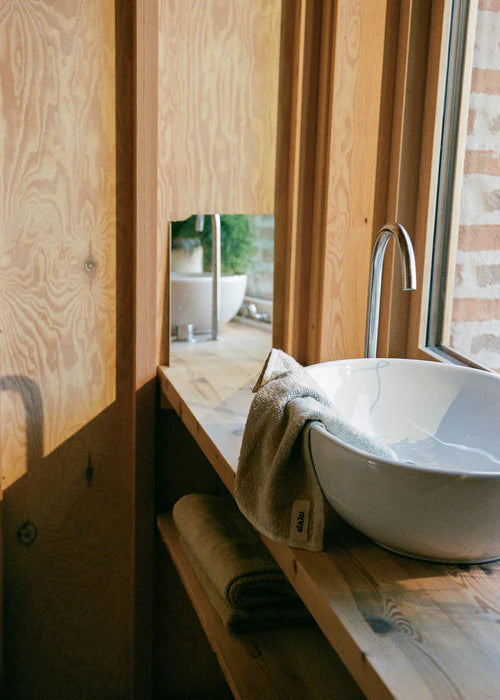 En hvid håndvask står på en træhylde med et gråt håndklæde ("daju") dækket over kanten. Vandhanen står over den fyldte vask, mens lys skinner gennem et vindue og reflekteres i overfladen.