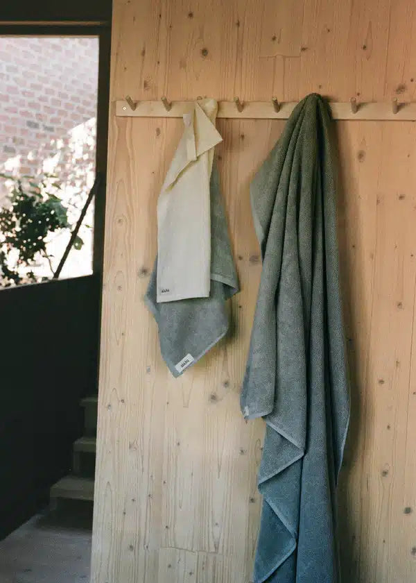 Håndklæder hænger på en knagerække monteret på en lys trævæg. Til venstre ses et vindue med grønt udenfor, og lidt af en trappe.
