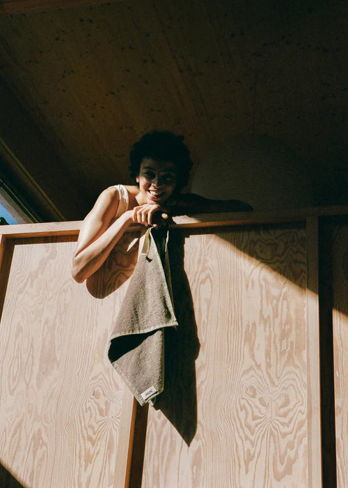 En kvinde smiler, mens hun læner sig over en træstruktur. Et gråt håndklæde med mærket "MUUD" hænger fra strukturen, og sollys lyser op i baggrunden.