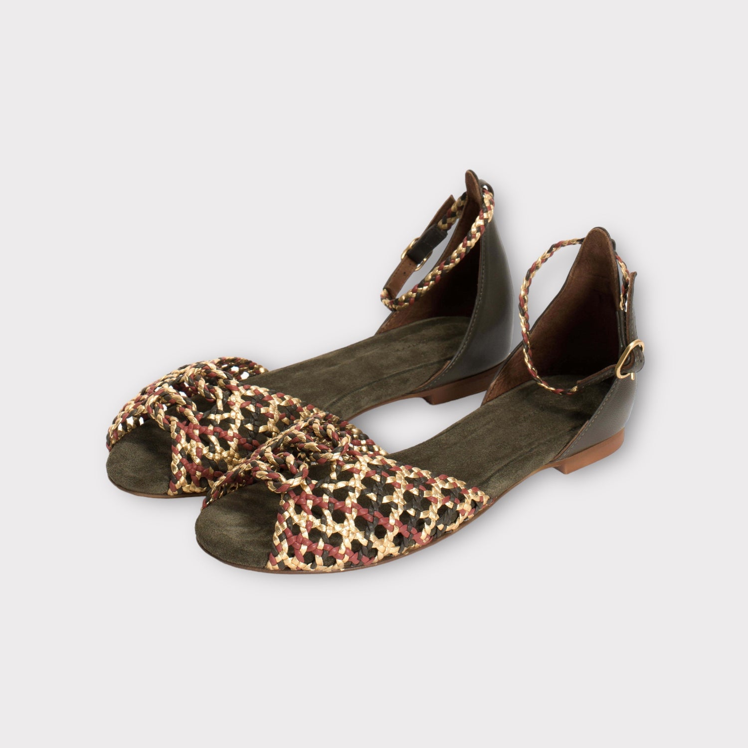 Et par flade sandaler med ankelrem ligger på en ensfarvet baggrund. Sandalerne har grønt ruskind og et flettet design i guld, rød og sort over tæerne.