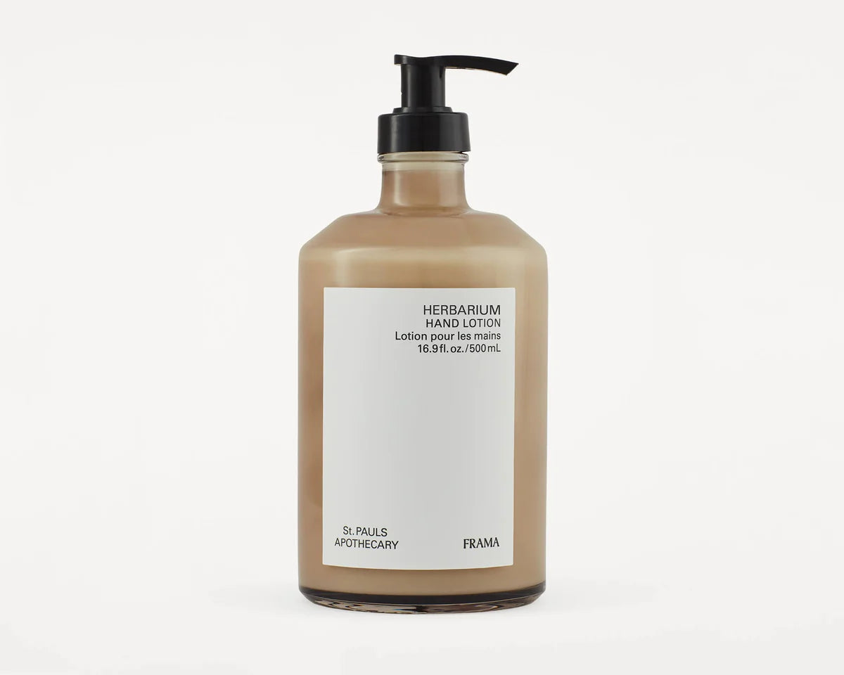 En brun glasflaske med sort pumpe indeholder "HERBARIUM HAND LOTION. Lotion pour les mains. 16.9 fl. oz./500mL". "St. PAULS APOTHECARY" og "FRAMA" står på etiketten. Flaskan står på en hvid baggrund.