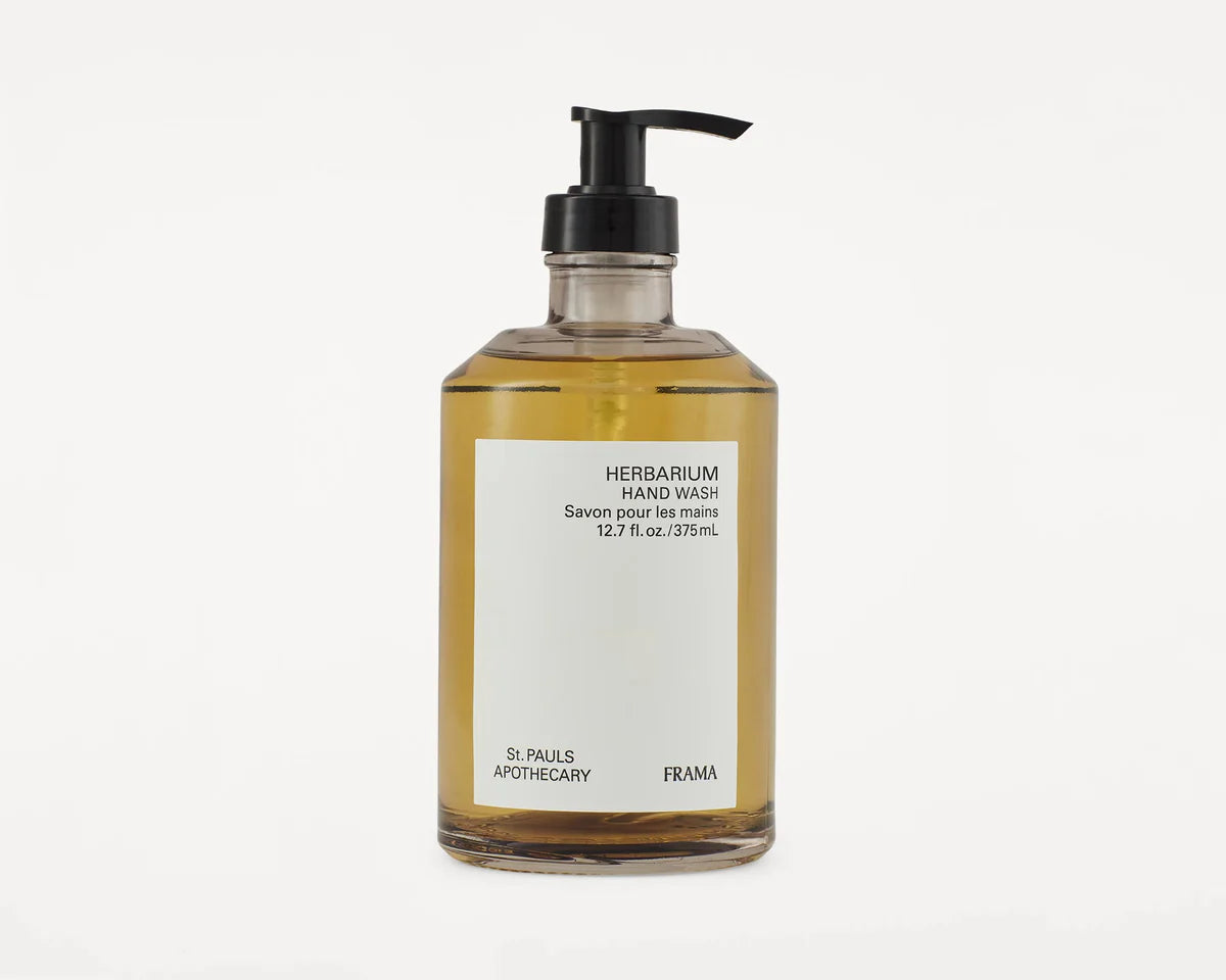 En klar glasflaske indeholder Herbarium håndsæbe, med etiket der angiver "HERBARIUM HAND WASH Savon pour les mains 12.7 fl. oz./375mL St. PAULS APOTHECARY FRAMA." Den har en sort pumpe og står på en hvid baggrund.