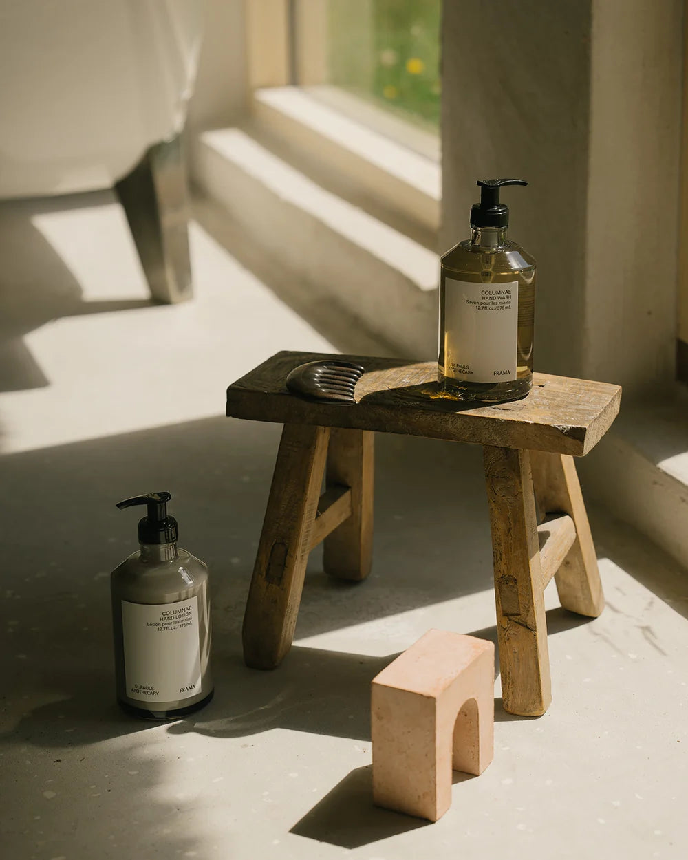 To flasker med pumpehoveder (COLUMNAE håndlotion og håndvask, 12.7 fl.oz./375 ml, ST. PAULS APOTHECARY, FRAMA) og en kam står på en lille træskammel i et badeværelse.