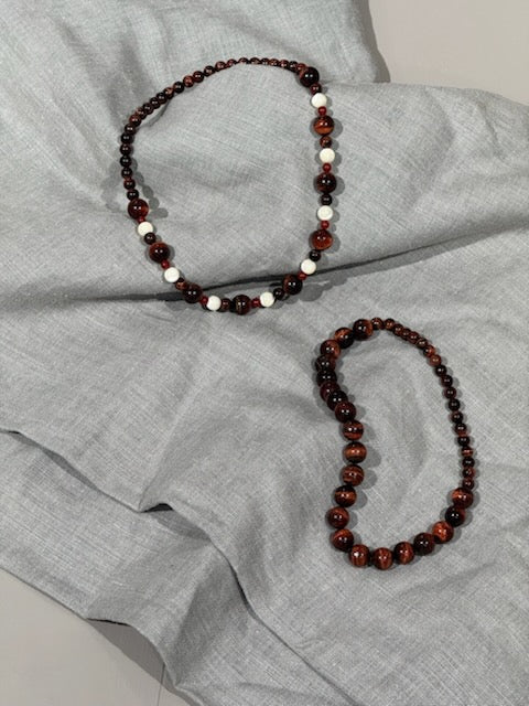 To halskæder, den ene med brune og hvide perler, den anden med kun brune perler, ligger på en lys grå stofoverflade.