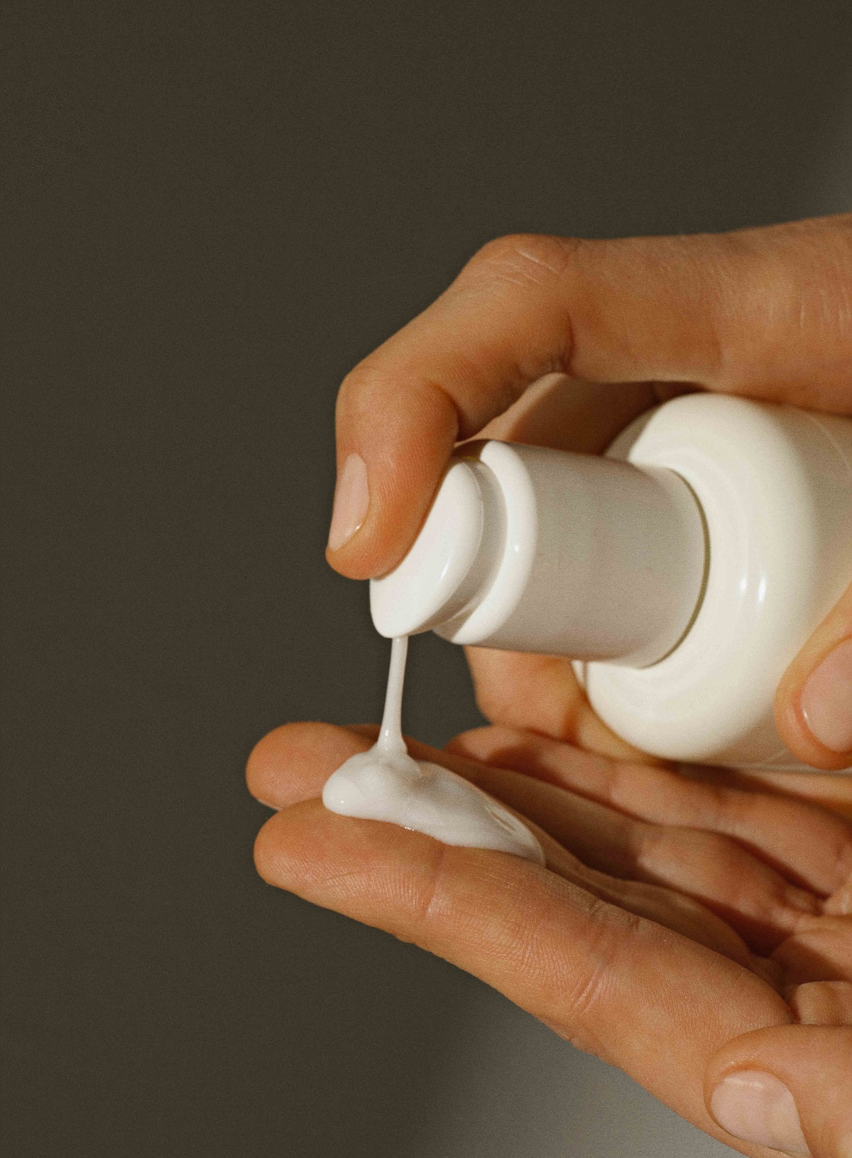 En hånd pumper hvid lotion fra en hvid flaske ned i en anden hånds håndflade mod en mørkegrå baggrund.