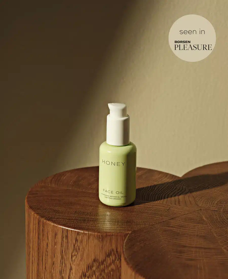 En grøn flaske "HONEY FACE OIL" står på en træstub. En cirkel med teksten "seen in BØRSEN PLEASURE" vises øverst til højre. Væggen bagved er lys beige.