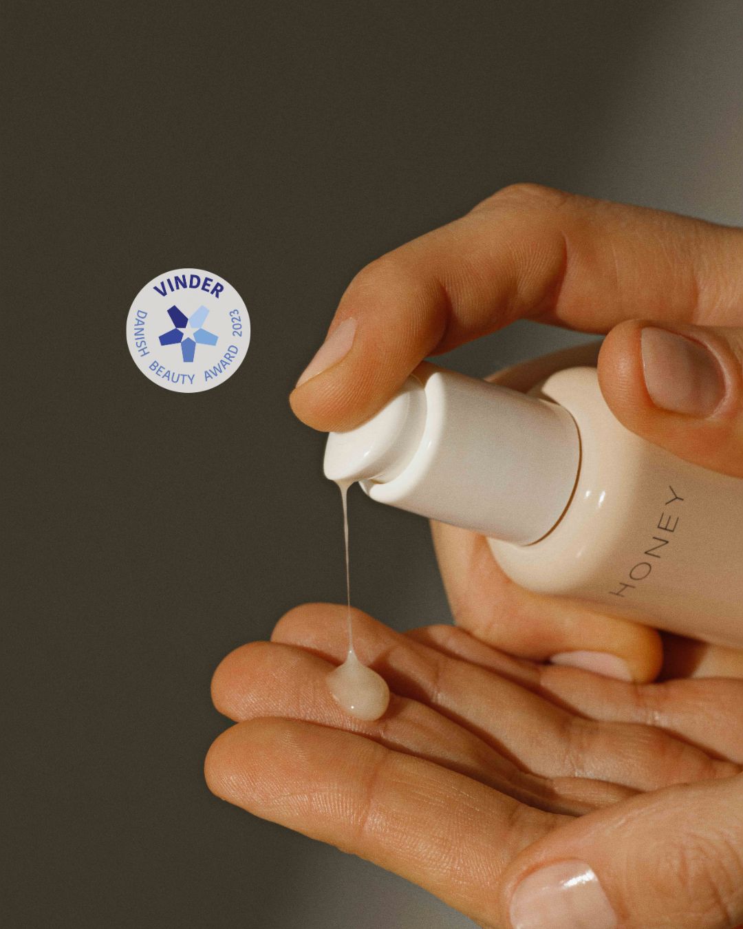 En hånd pumper en creme med navnet "HONEY" ud i en anden hånd. I baggrunden ses et "Vinder Danish Beauty Award 2023" mærke.