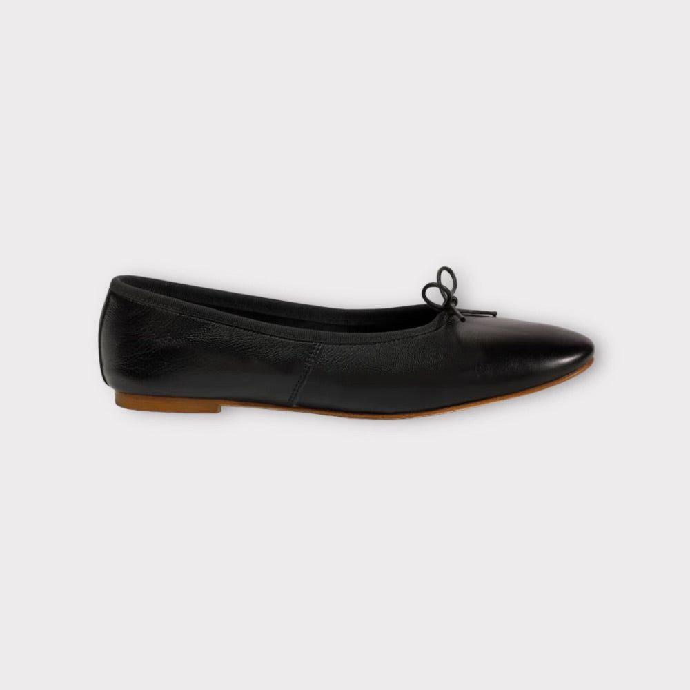 En sort ballerina-sko hviler på en ren, gråhvid baggrund. Skoen har en lille sløjfe på tåen og en lav, brun hæl.