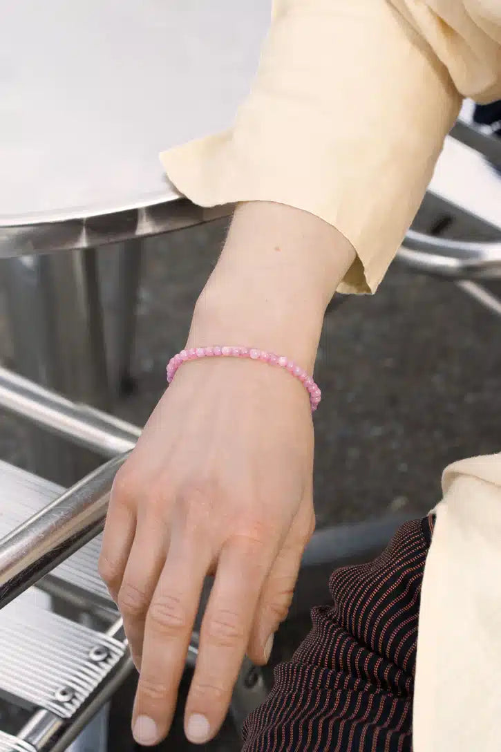 En lyserød perlearmbånd sidder på en persons håndled. Personen har en lys gul skjorte på og sidder ved et sølvfarvet bord.