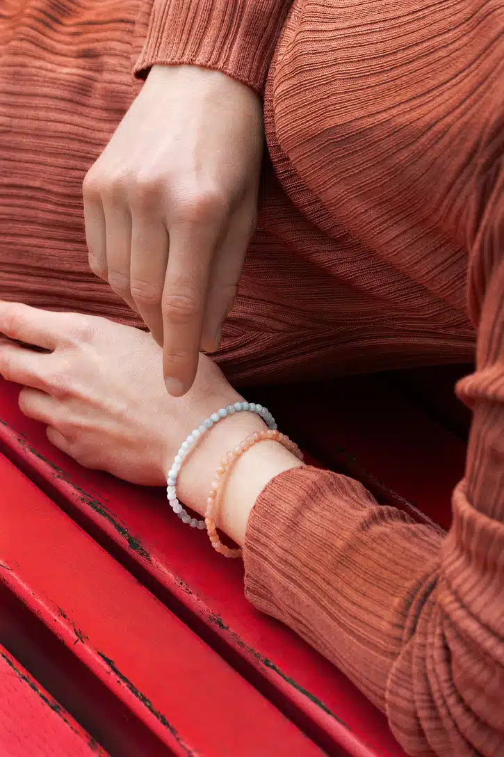 En hånd, iført to perlearmbånd, hviler på et rødt træunderlag, med en anden hånd let hævet over håndleddet i et terracottafarvet striktøj.