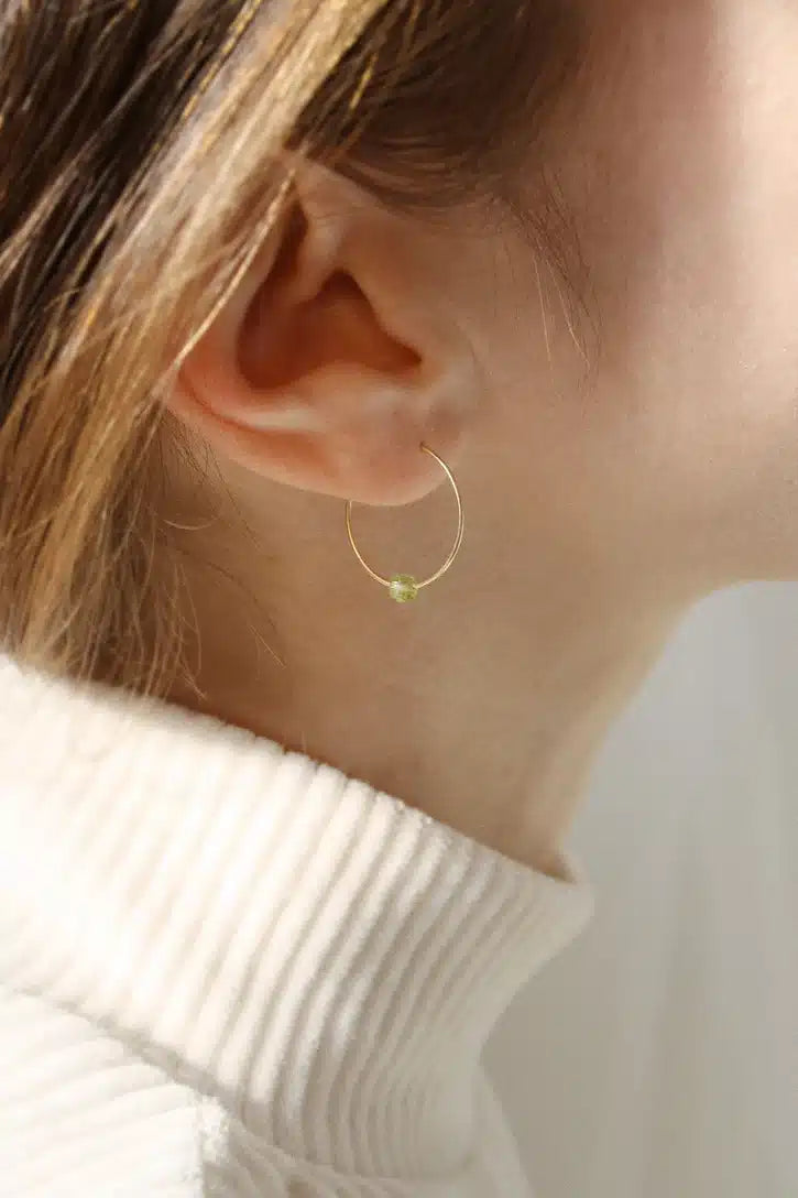 Et kvindeligt øre bærer en guldfarvet, cirkelformet ørering med en grøn perle. Øreringen hænger ned mod en hvid rullekrave.