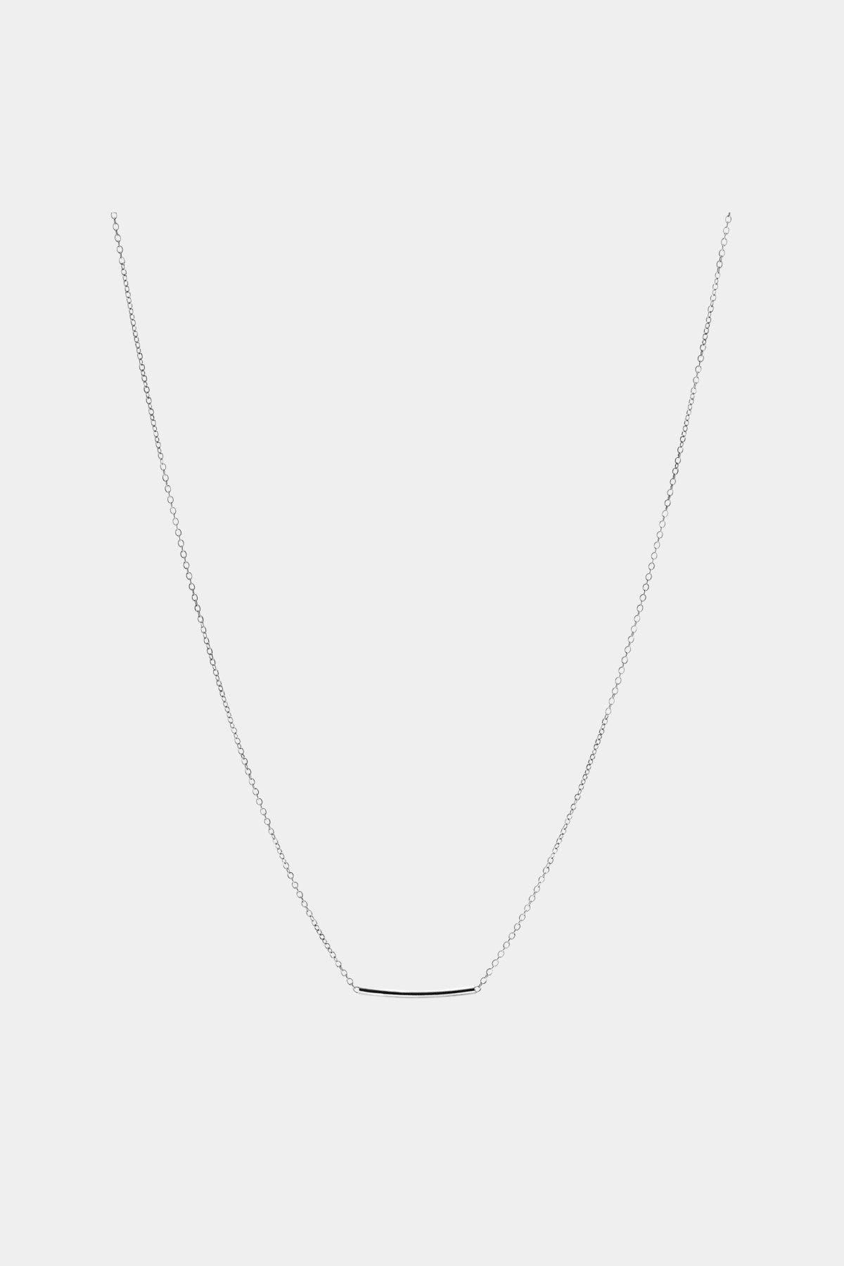 En sølv halskæde med et tyndt, horisontalt stykke som vedhæng hænger mod en ensfarvet, lysegrå baggrund.