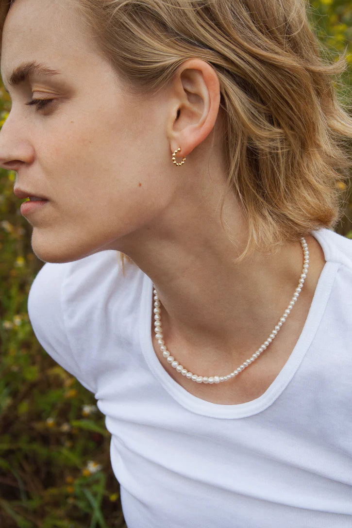 En ung kvinde med blondt hår bærer perlehalskæde og guldøreringe. Hun kigger til venstre, poserer i en hvid T-shirt, med en naturlig baggrund.