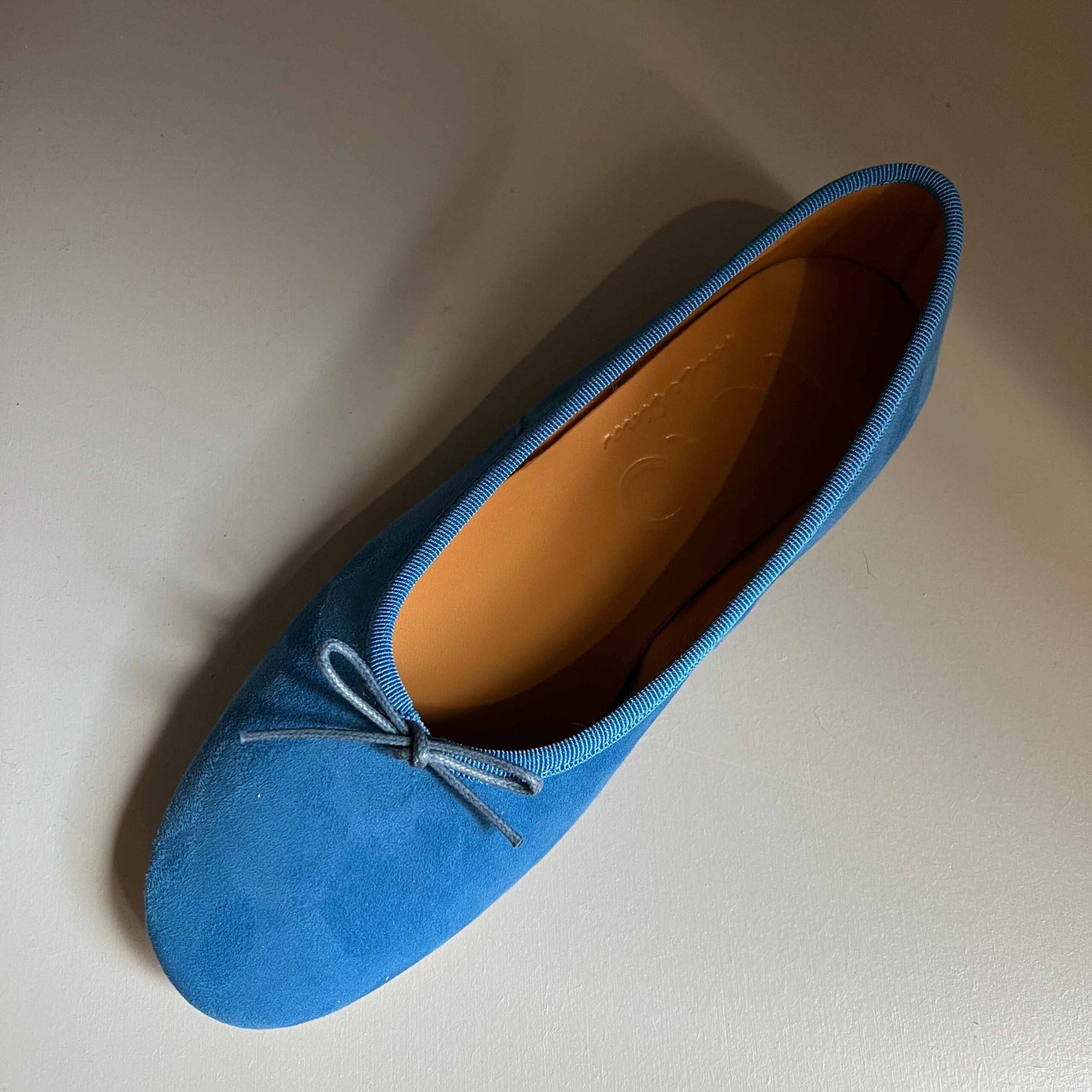 En enkelt lyseblå ruskindssko ligger på en hvid overflade. Skoen er en ballerina model med en lille sløjfe. Indersiden er orange.