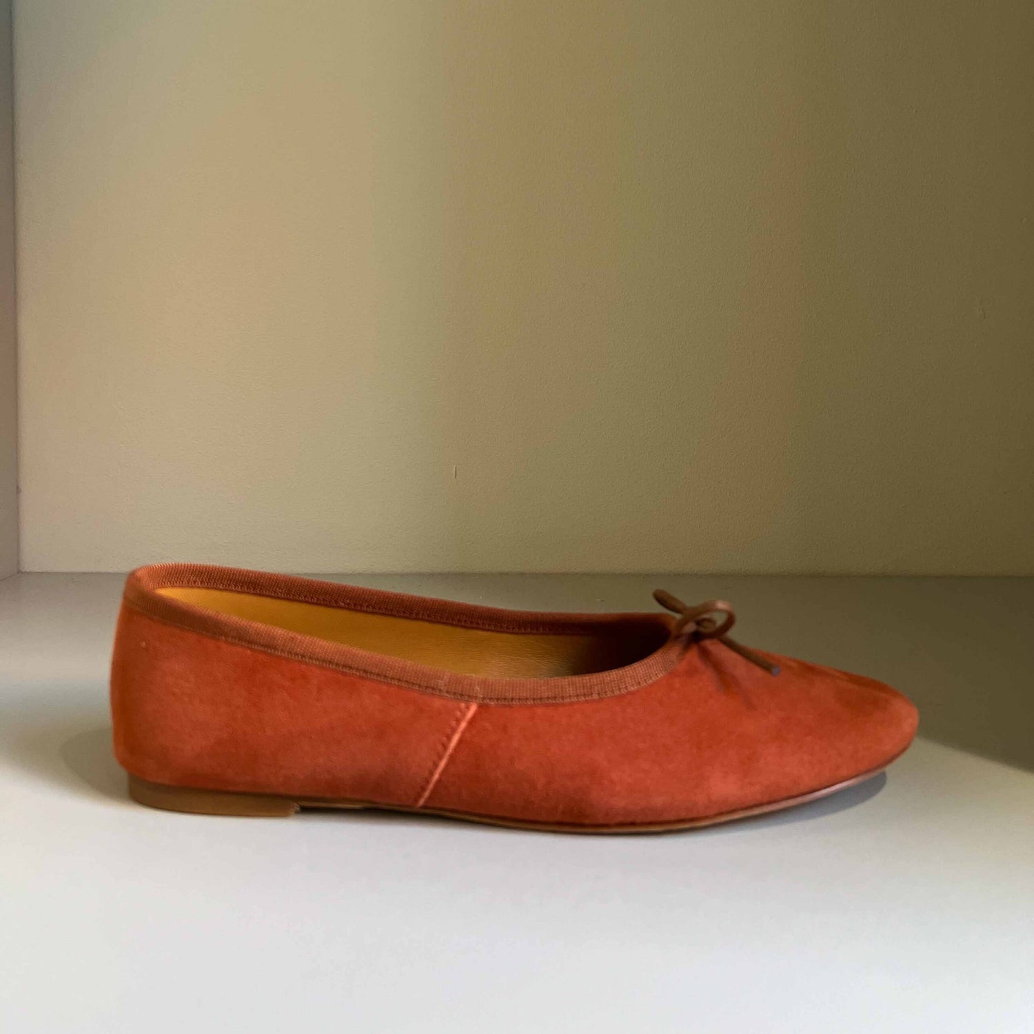 En orange ballerina sko ligger på en hvid overflade mod en beige baggrund. Skoen har en lille sløjfe på snuden.