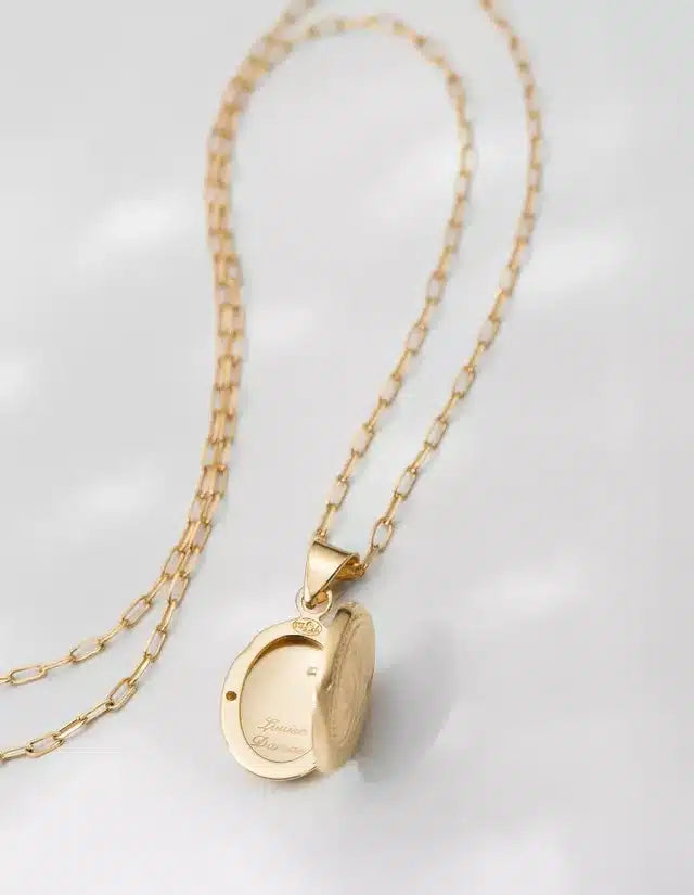 Et guldfarvet medaljonhalskæde hænger på en hvid overflade. Medaljonen er åben og viser inskriptionen "Louise Damas" indeni.